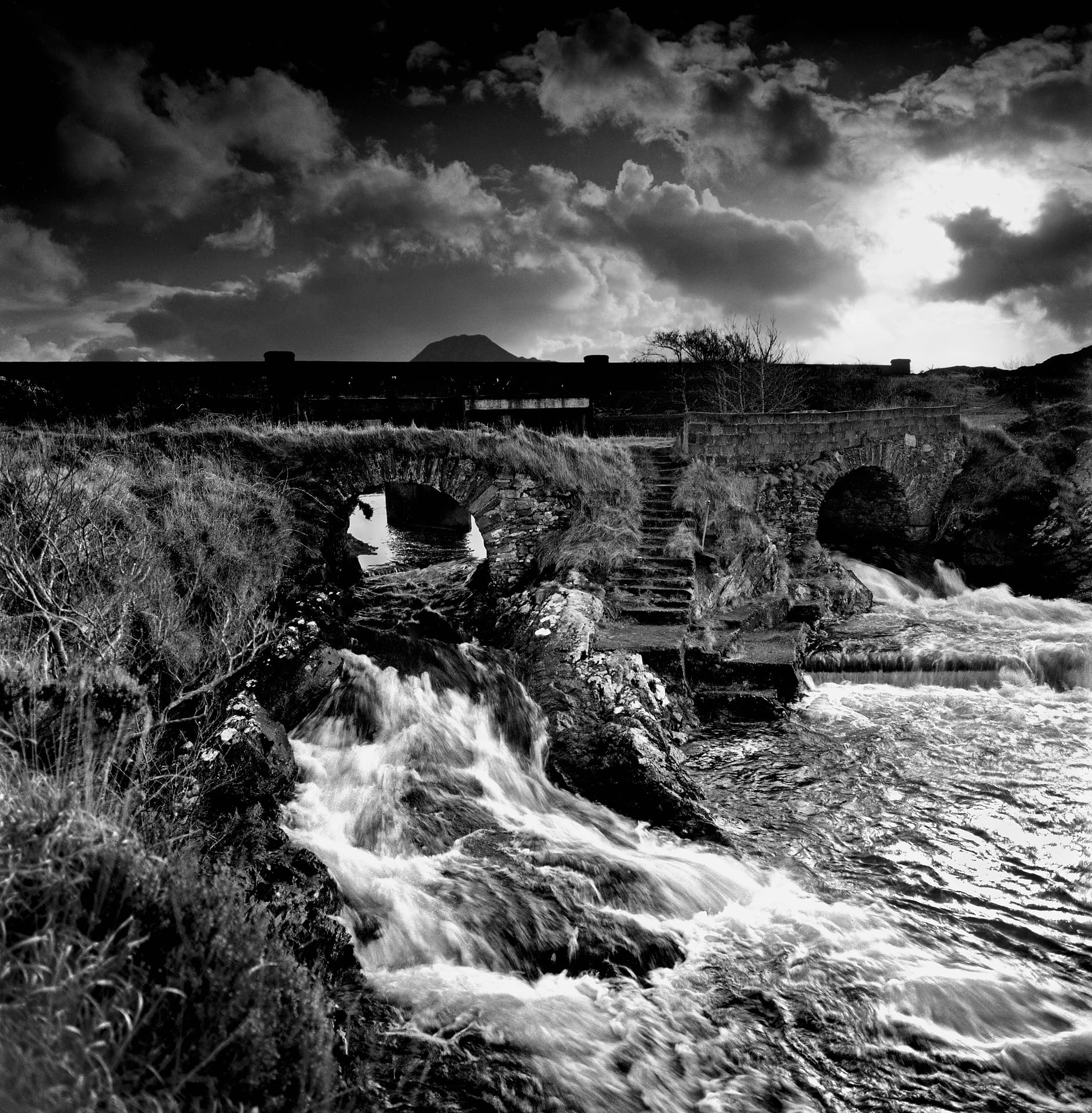 Water rushing under a stone arch bridge below a dark cloud-filled sky, moorland grass on both banks